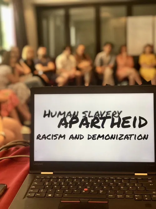 Ein aufgeklappter Laptop mit der Folienaufschrift "Human Slavery, Apartheid, Racism and Demonization" steht auf einem Tisch, dahinter sind unscharf Personen in einem Stuhlreis zu sehen.
