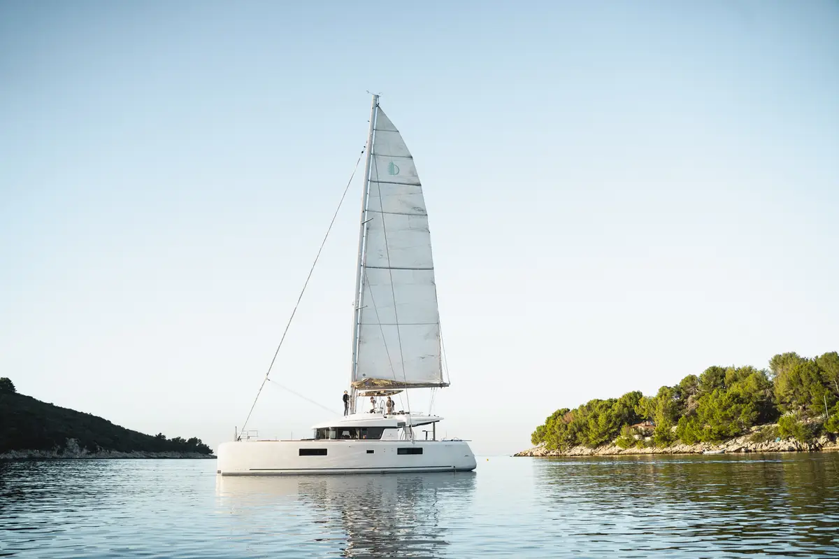 Catamaran under sail near a green, rocky coastline